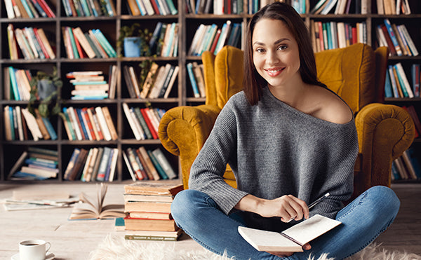 An author at ease in a reading chair, notebook in hand, surrounded by books